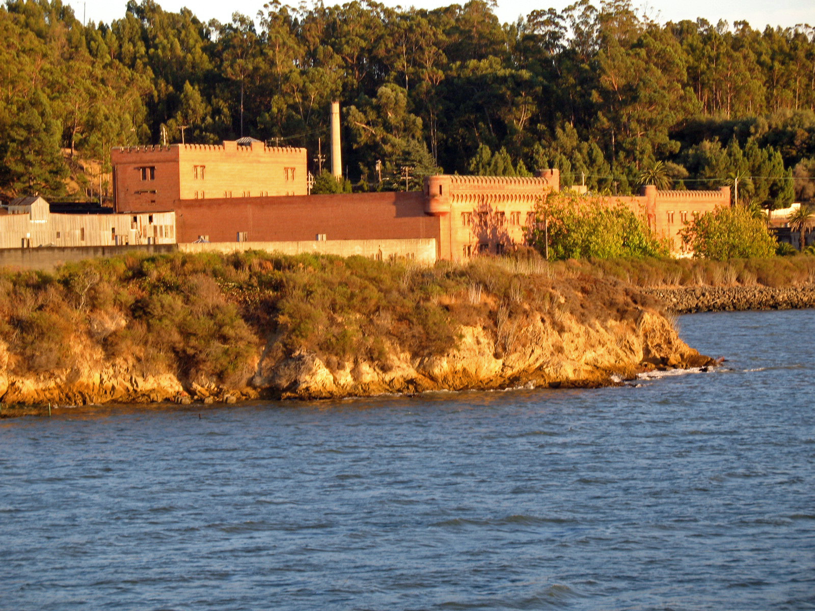 Winehaven’s brick cellars and smokestack seen across the water from the northwest, with wooded hills behind